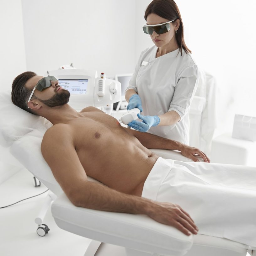 Young master with goggles conducts laser epilation to bearded man client lying on couch in clinic